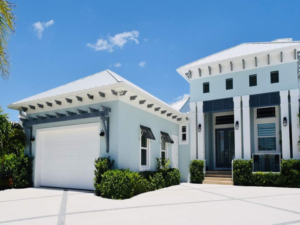 dark blue bahama shutters on a light blue hosue with lush green shrubs