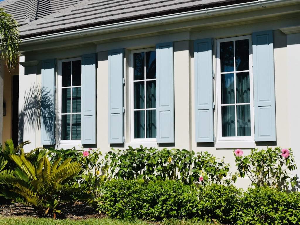 light blue colonial shutters on an off white house with hibiscus shrubs under the windows